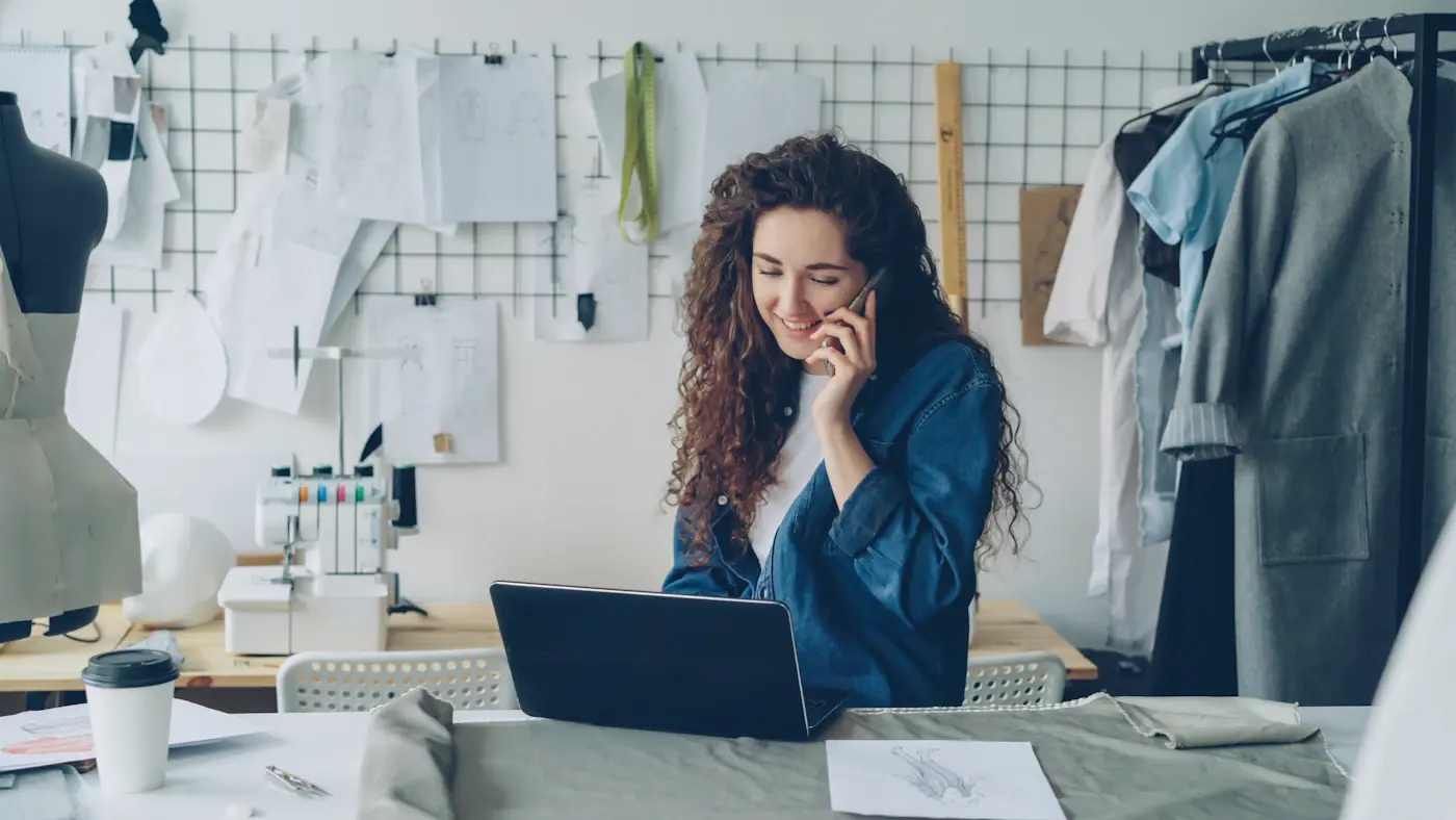 A designer on the phone at a laptop in a studio full of fabric